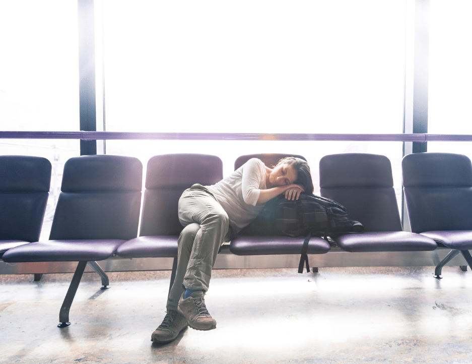 Woman asleep in an airport, lying on bench seats, resting her head on her bag.