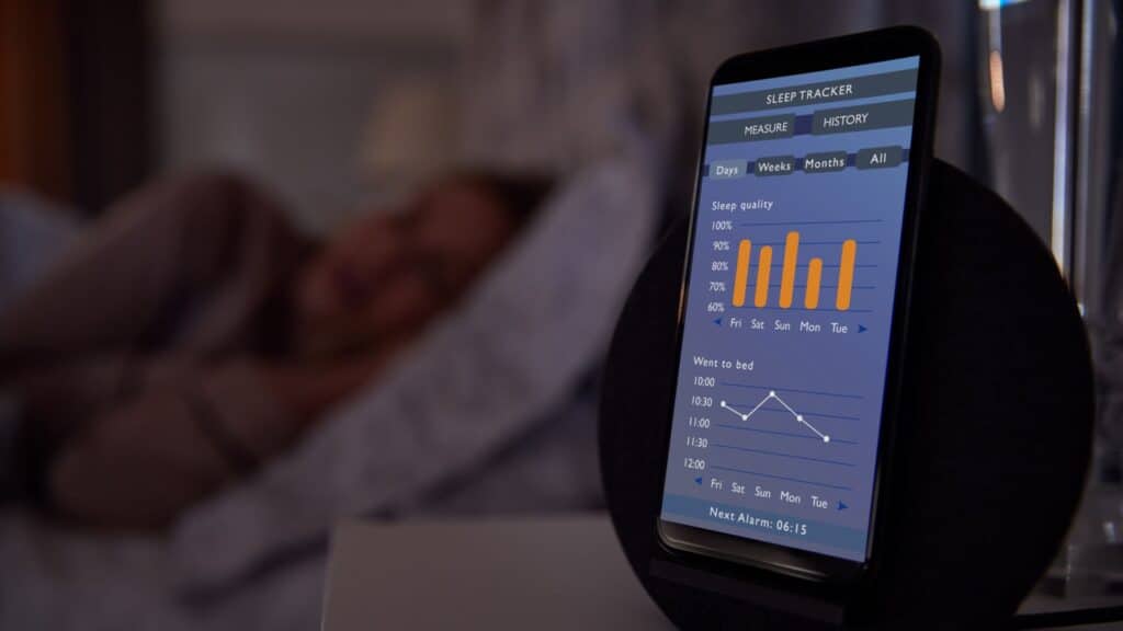 Photo of a a woman asleep in bed, with her smartphone in the foreground. The smartphone has images of the sleep data it is tracking while she sleeps.