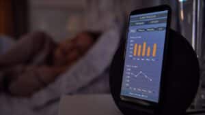 Photo of a a woman asleep in bed, with her smartphone in the foreground. The smartphone has images of the sleep data it is tracking while she sleeps.
