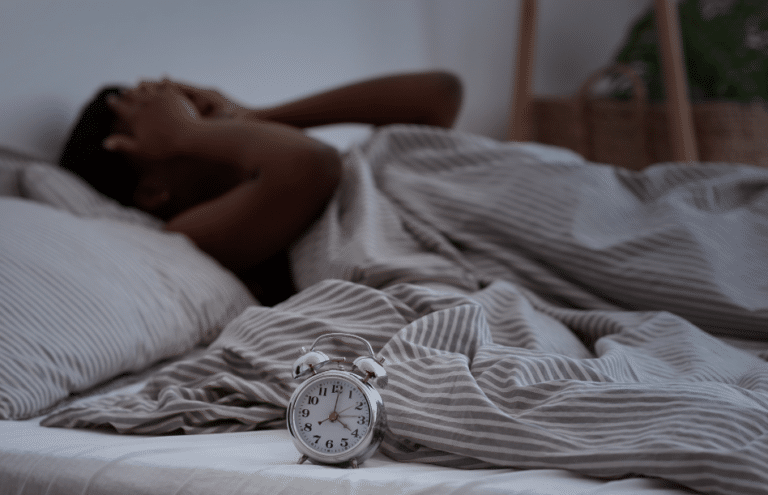 Photo of a person lying awake in bed in the dark, with their hands on their face, looking stressed. Clock in the foreground.